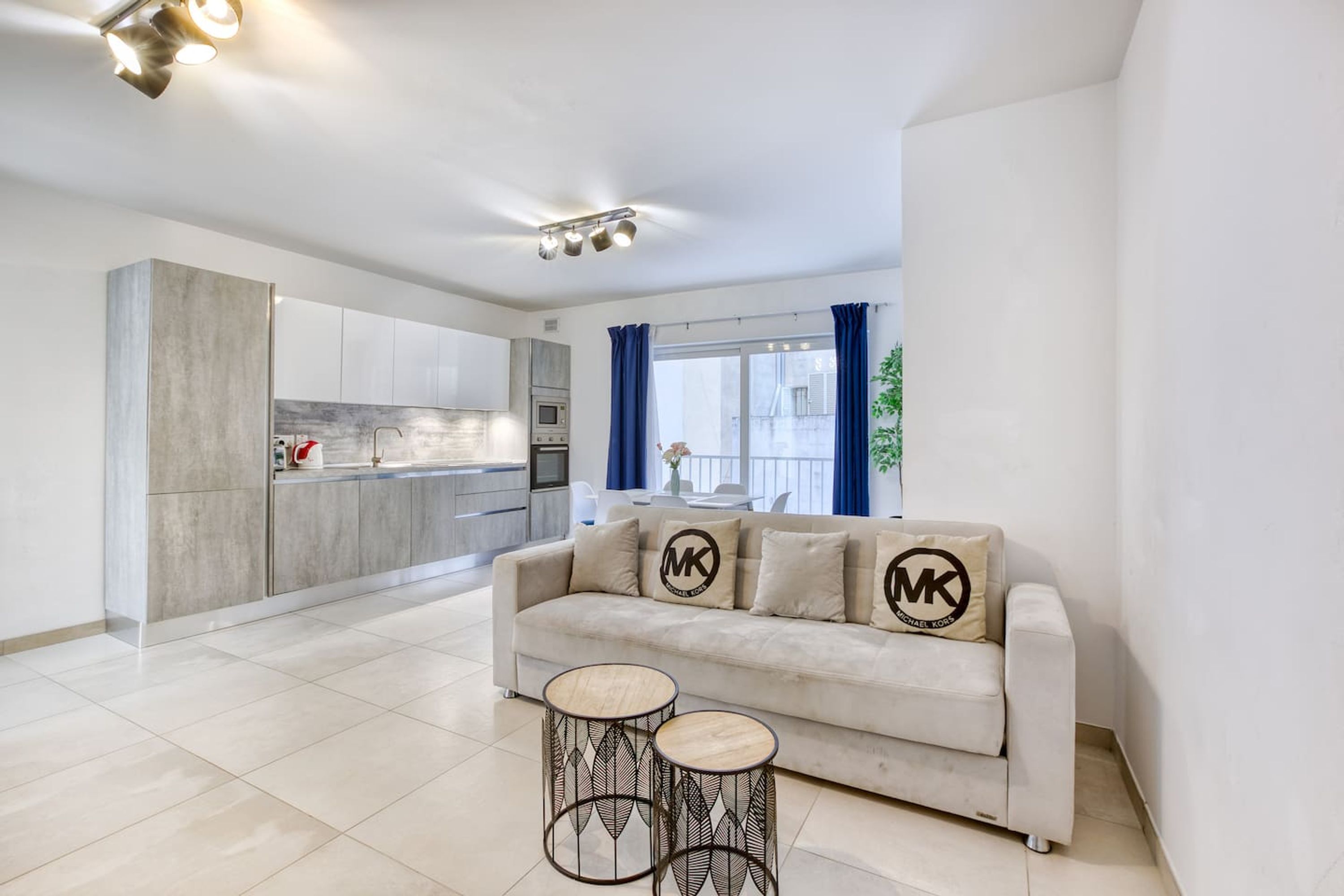 Una sala de estar moderna con un sofá, dos mesitas auxiliares y una cocina al fondo.