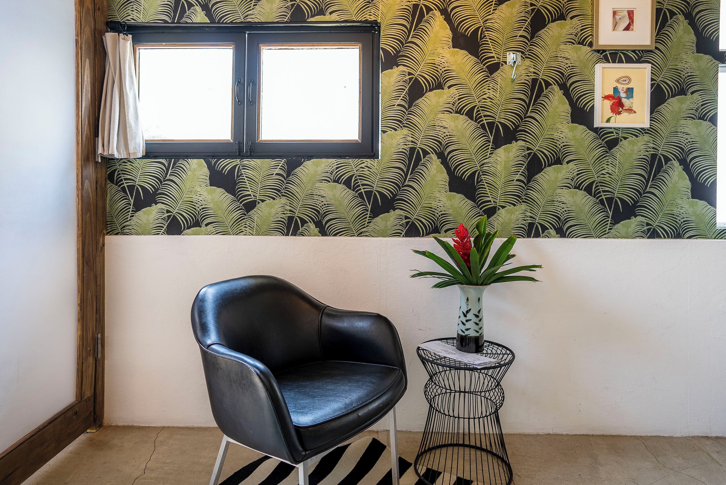 Una habitación con una butaca de cuero negro, una mesita redonda con un florero, una ventana con cortinas y una pared con un motivo de hojas.