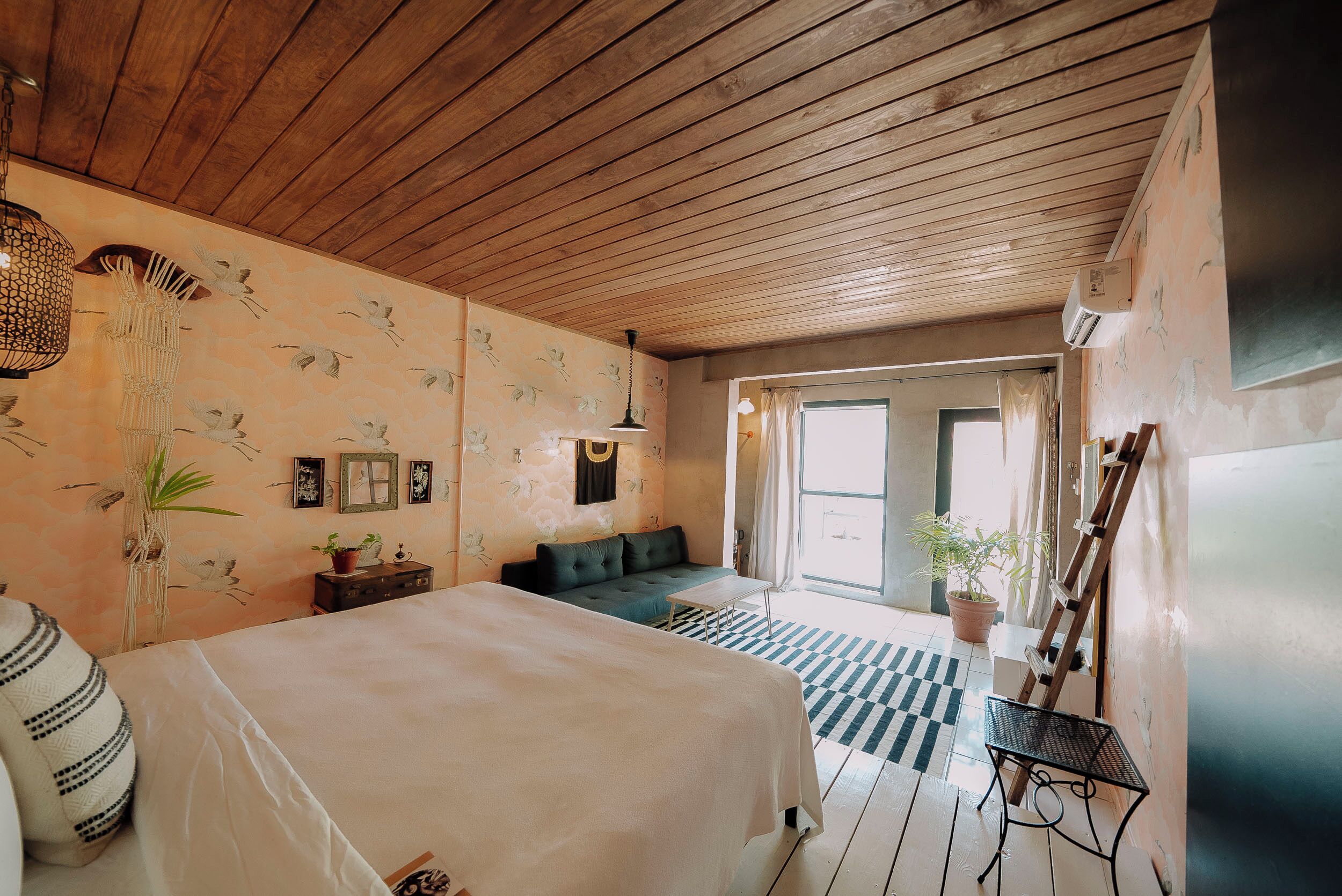A bedroom with a wooden ceiling, a bed, a sofa, a table, and a view of the outside.