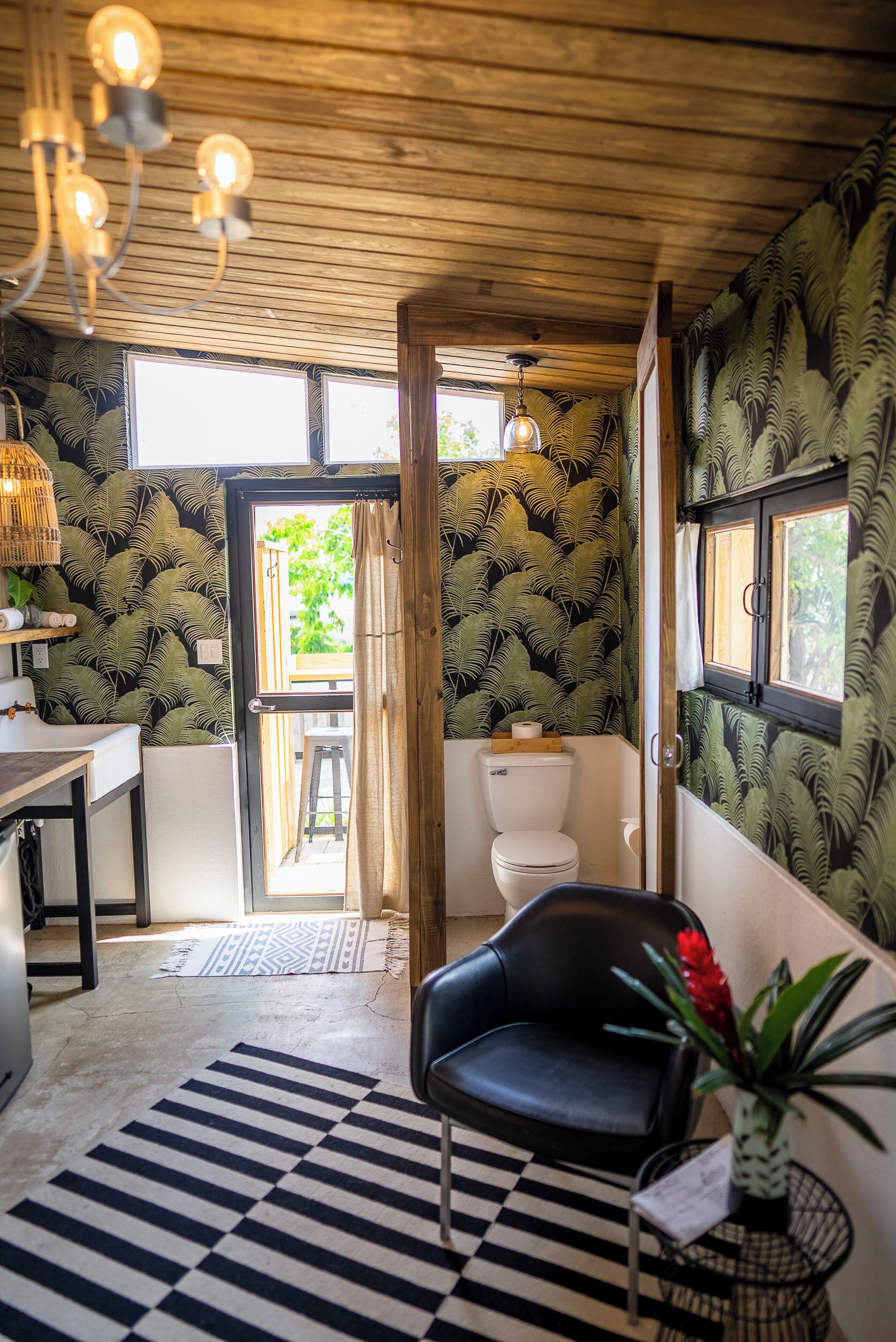 A room with a wooden ceiling, a black chair, a striped rug, a sink, and a toilet with a fern wallpaper.