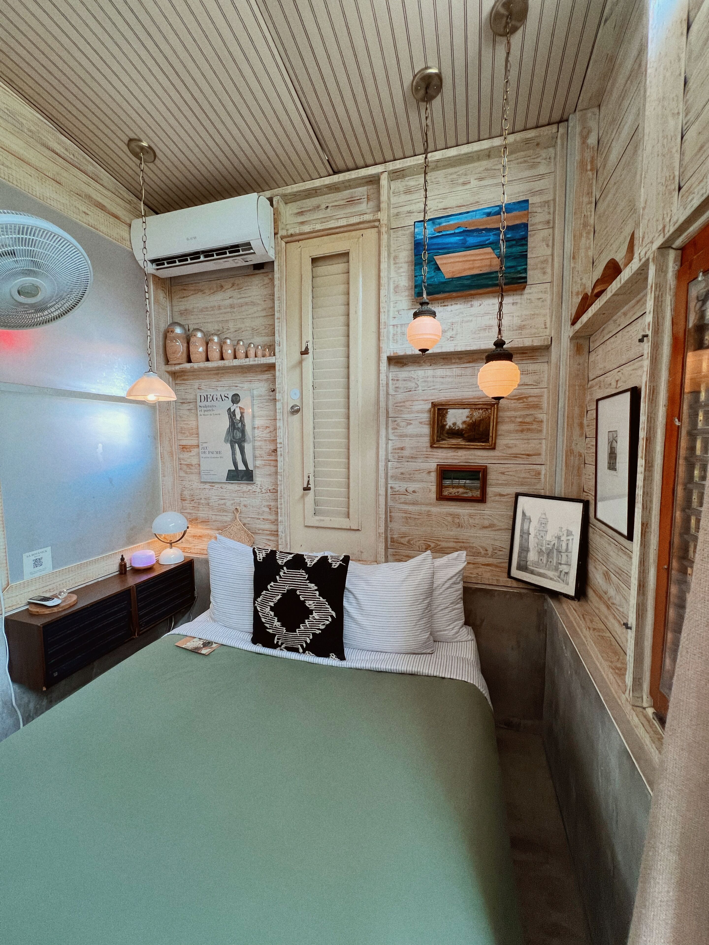 A bedroom with a bed, bedside table, lamp, wall-mounted air conditioner, and a ceiling fan.