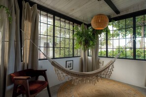 A hammock in a room with large windows, a chair, and a woven rug. - Casa Botanica (San Juan)