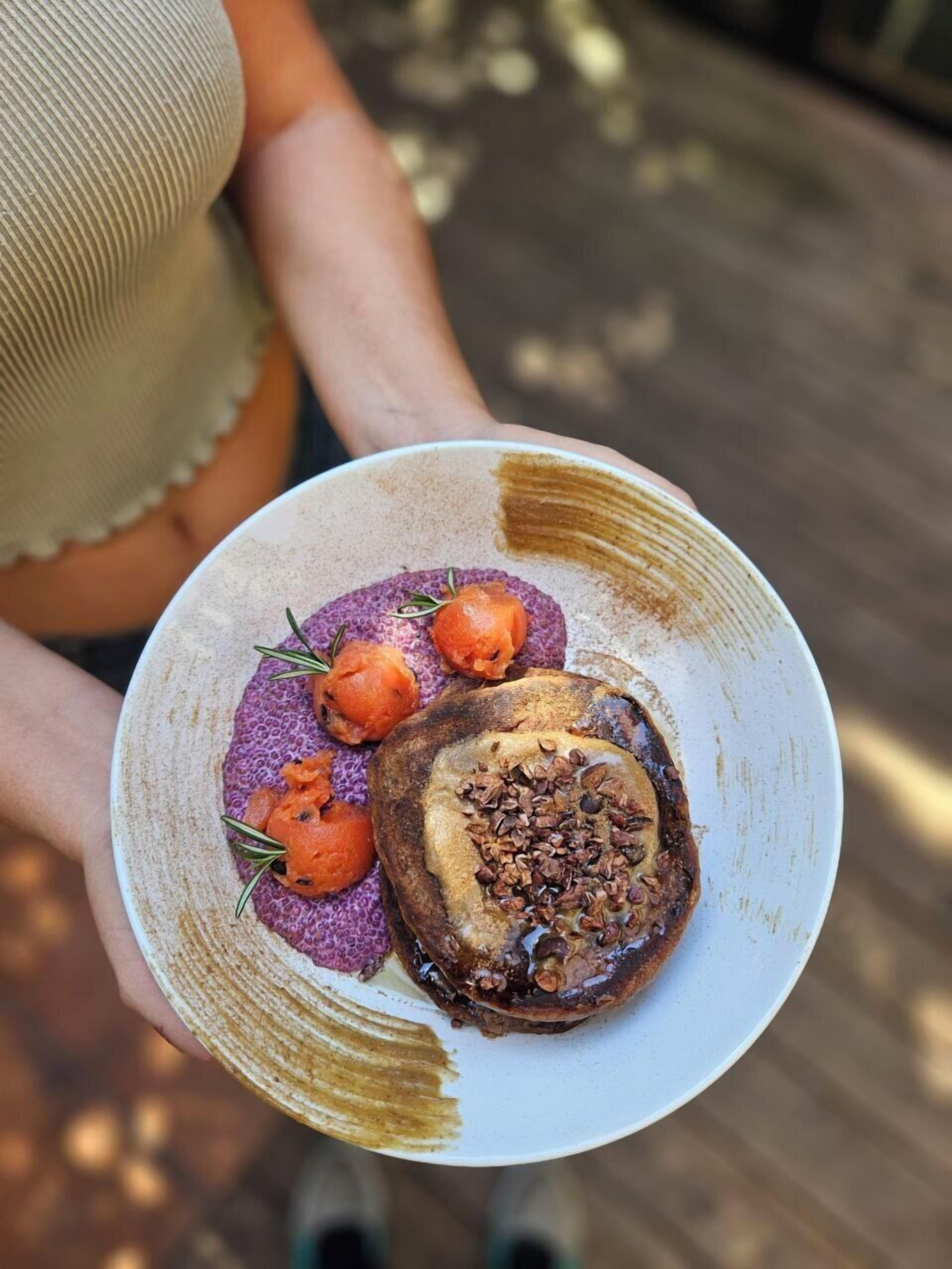 A person holding a plate with a dessert that includes a chocolate cake, a purple puree, and orange fruit garnishes.