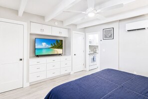 Une chambre à coucher avec un lit, un téléviseur posé sur un meuble, un ventilateur de plafond et un coin cuisine.