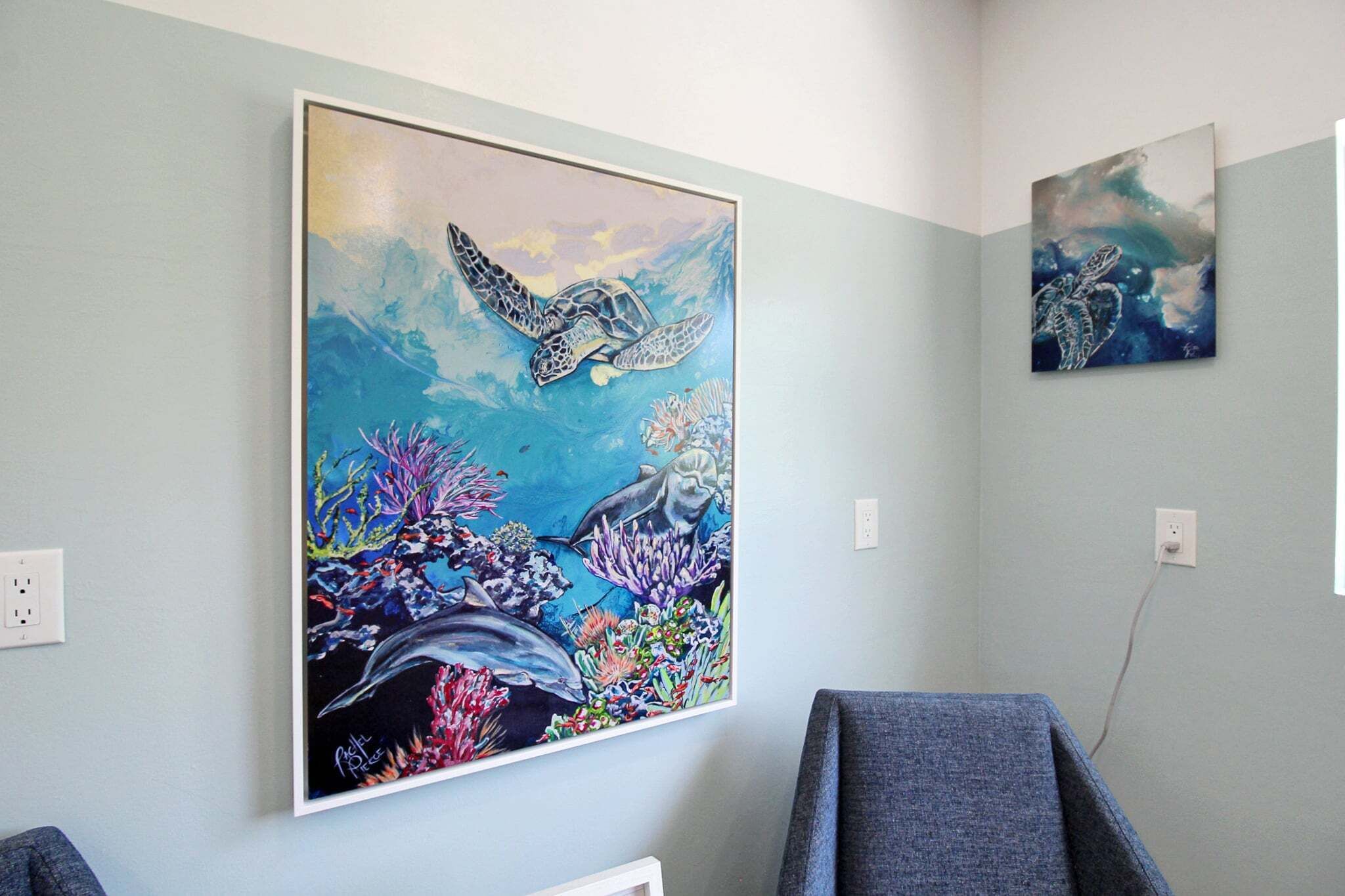 A room with a large painting of a sea turtle and coral reef on the wall, a gray chair, and a white electrical outlet.