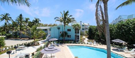 A pool area with lounge chairs, umbrellas, and a building with multiple floors and balconies.