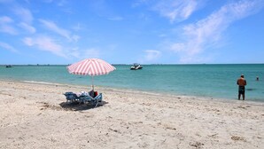 A beach with a striped umbrella, lounge chairs, and a person standing by the water. - Relaxing 1BR Sanibel Hideaway - Colony Inn 13 (Sanibel)