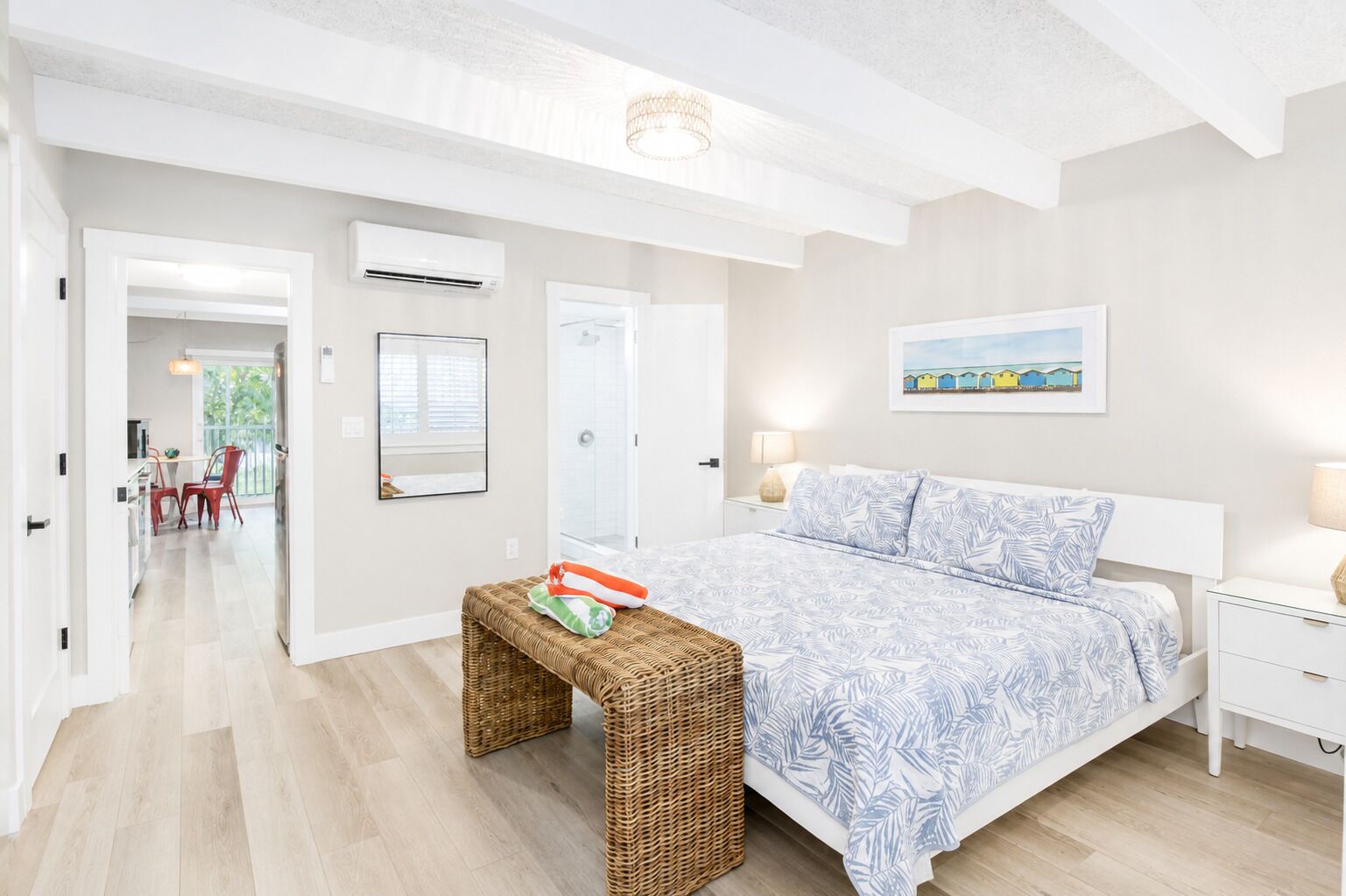 Une chambre à coucher avec un lit, une table de chevet et un banc en osier. Une porte donne accès à une salle de bain et à un couloir.