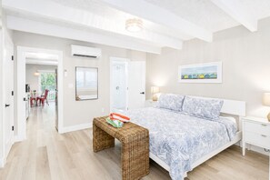 Een slaapkamer met een bed, nachtkastje en een rieten bankje. Er is een deur naar een badkamer en een gang.