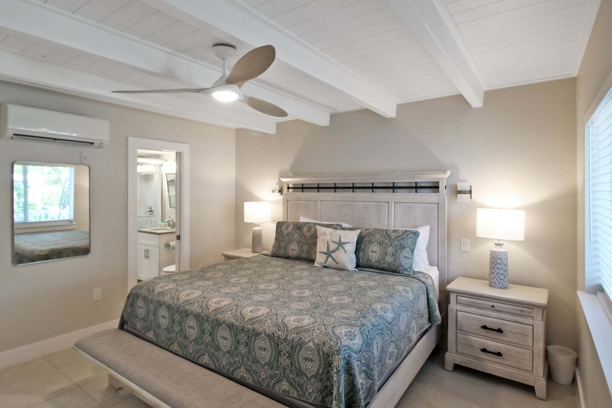 A bedroom with a bed, a ceiling fan, a nightstand with a lamp, a bench, a window, and a mirror.