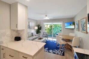 Een moderne keuken met witte kasten, een eethoek met een blauw gestreept vloerkleed en een tv waarop een strandscène te zien is.