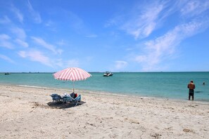 Ein Strand mit einem gestreiften Sonnenschirm, Liegestühlen und einer Person am Wasser.