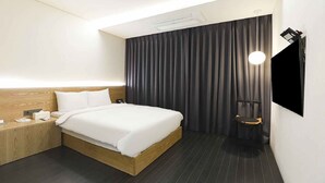 Une chambre d’hôtel moderne avec un lit en bois, une petite table de chevet, un téléviseur fixé au mur et un rideau foncé.