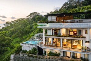 Una casa moderna de varios pisos con piscina, balcones y abundante vegetación.