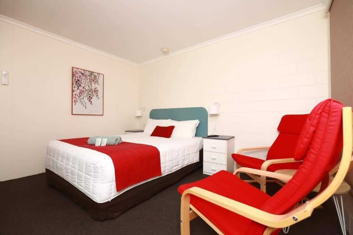 A hotel room with a bed, a nightstand, a chair, and a wall-mounted lamp.