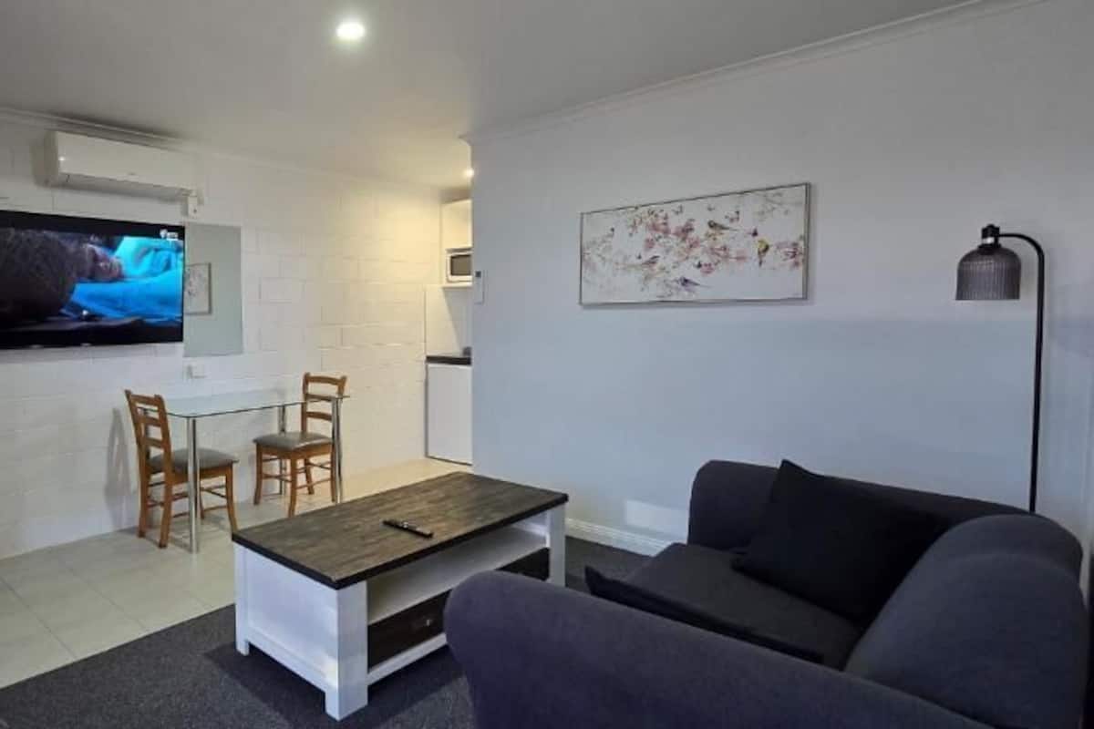 A living room with a sofa, a coffee table, and a TV mounted on the wall. There is a dining area with a table and chairs.