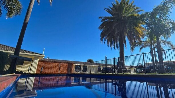 A swimming pool with a wooden fence, palm trees, and a clear blue sky. - Albatross Motor Inn (Lakes Entrance)