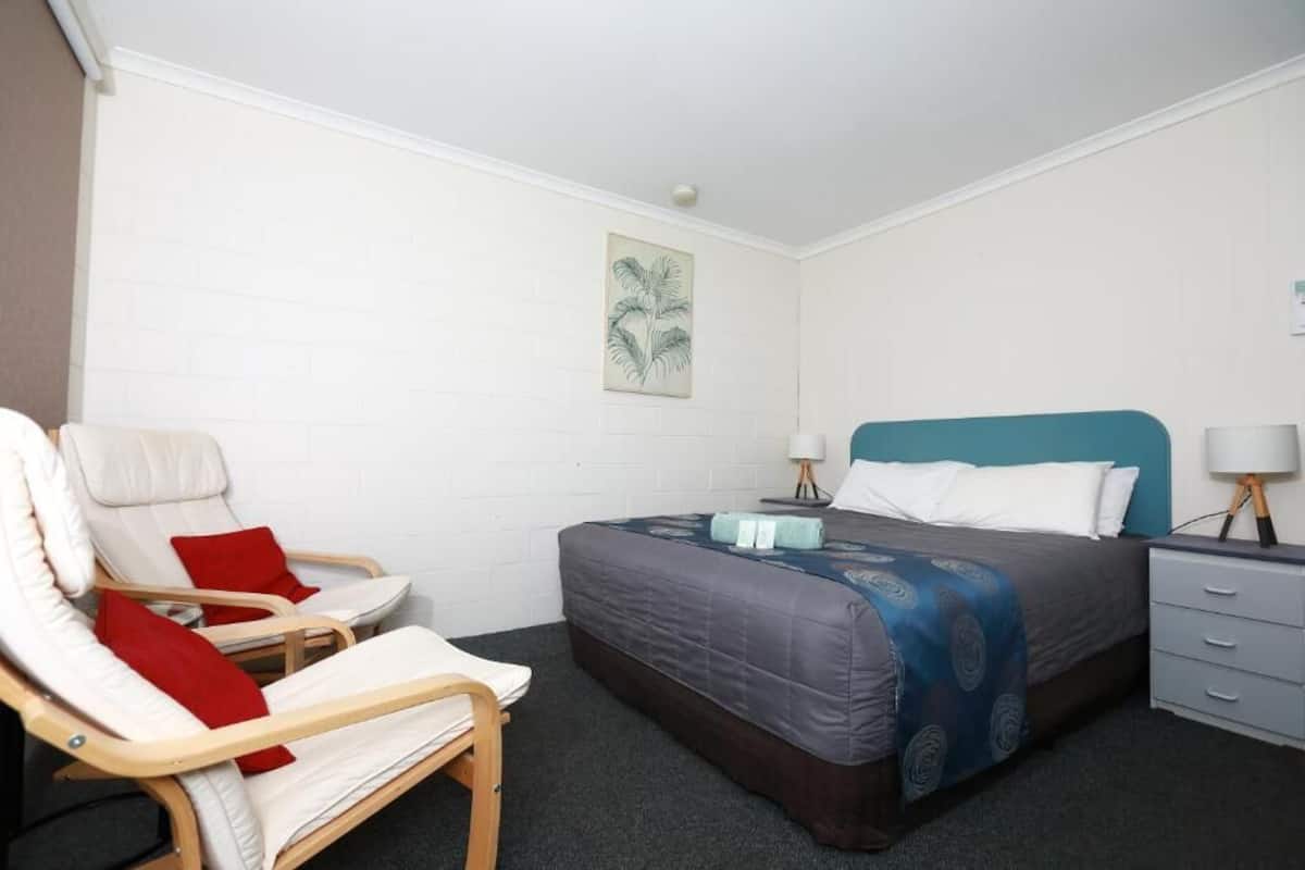 A single bed with a blue headboard, a bedside table with a lamp, a chair with red cushions, and a wall-mounted artwork of a palm tree.