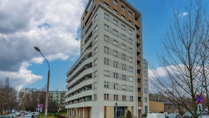En flervåningsbyggnad med bostäder, balkonger, en tydligt markerad entré och utsikt över gatan med parkerade bilar och en klar himmel.