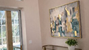 Un interior moderno con un ventanal que da a un balcón, una consola dorada con una planta en maceta y un cuadro abstracto en la pared.