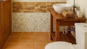 Ein Badezimmer mit einem Holzkosmetikschrank, einem weißen Waschbecken und einer Toilette. Eine Zierwand mit Steinfliesen und ein Gitterfenster über dem Waschbecken.
