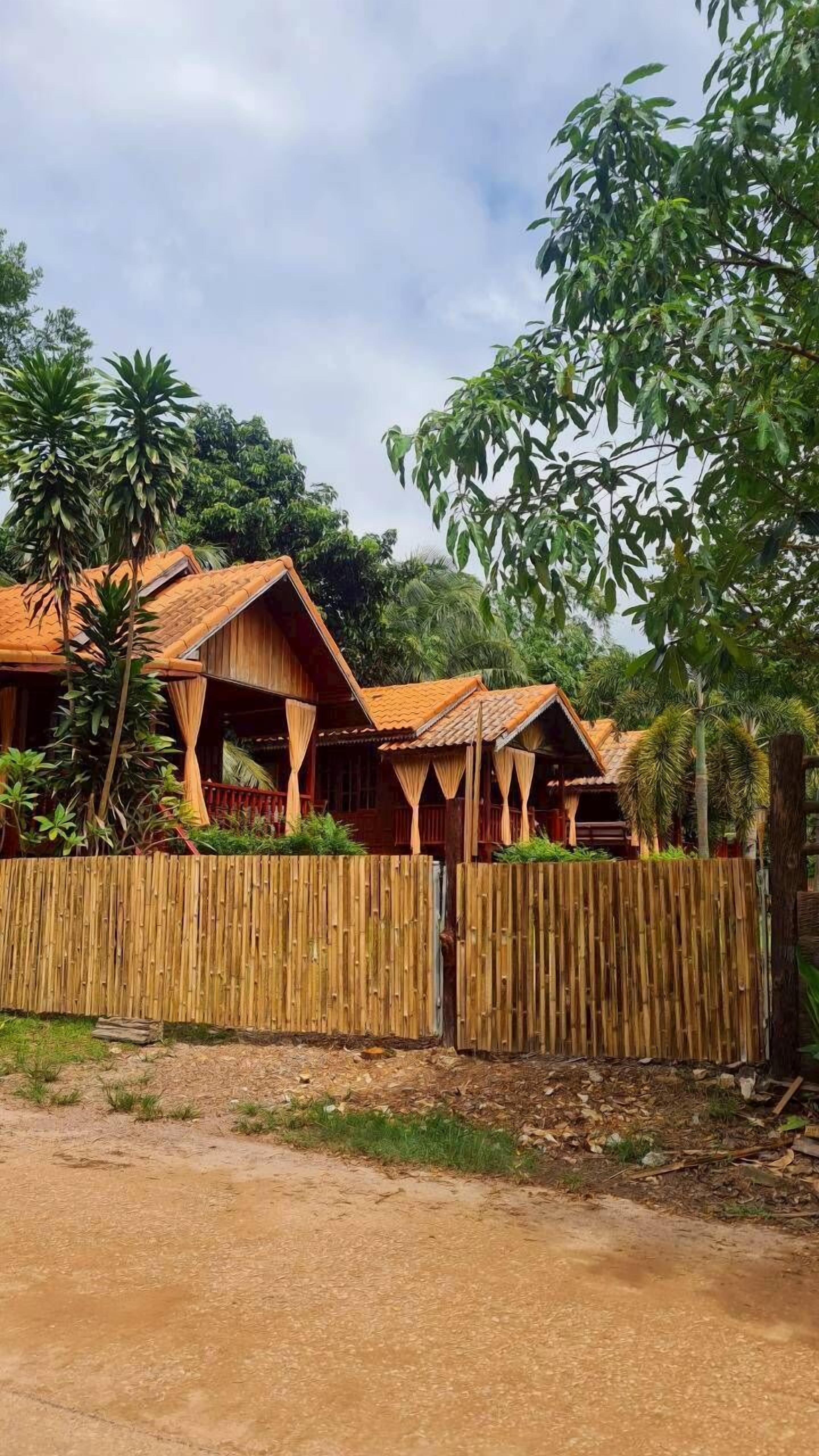 A row of wooden cabins with thatched roofs, surrounded by bamboo fencing and lush greenery.