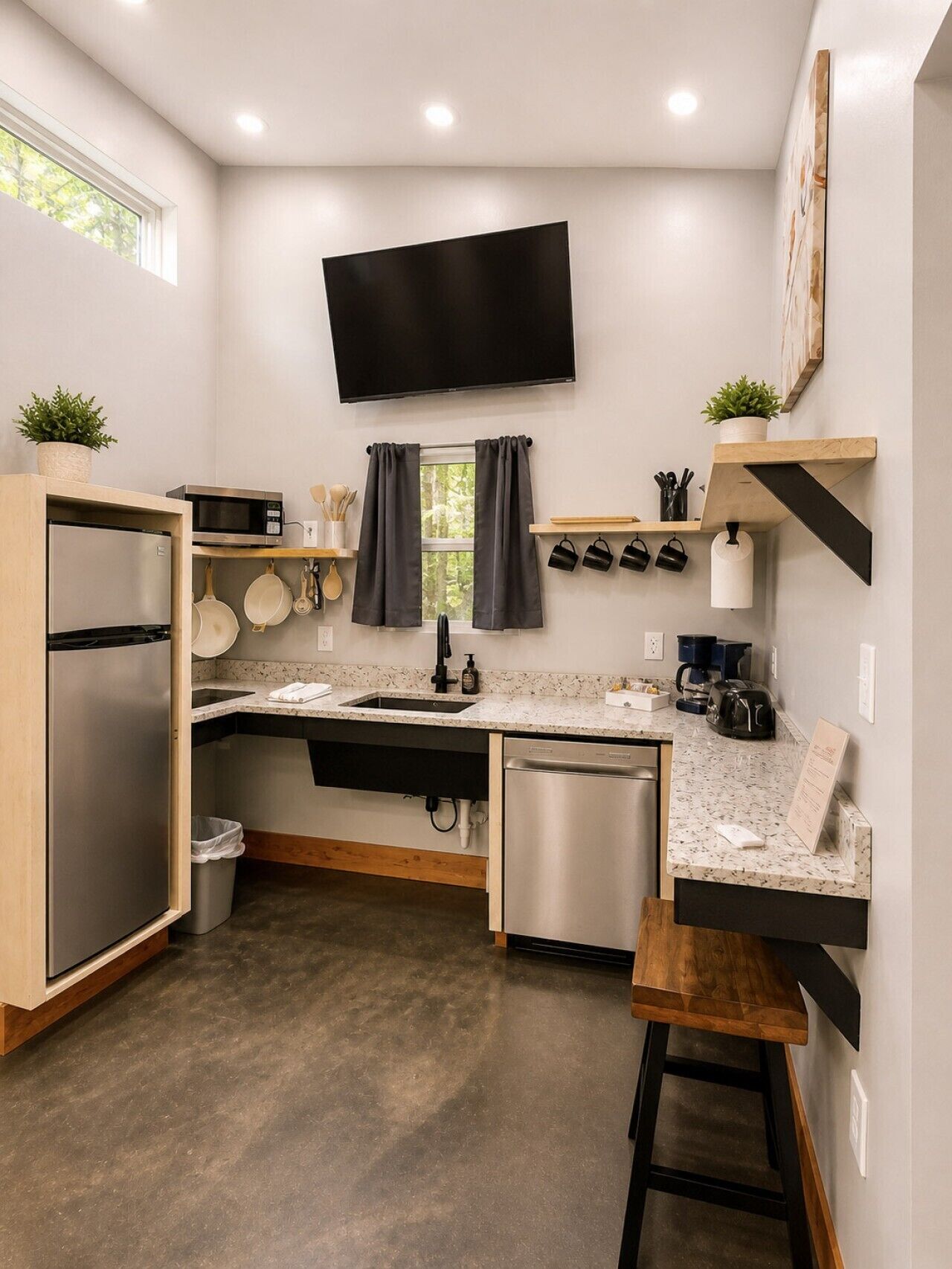 A compact kitchen with a microwave, stove, sink, and dishwasher. There's a flat-screen TV mounted above the counter, and a window with curtains.