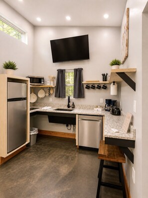 A compact kitchen with a microwave, stove, sink, and dishwasher. There's a flat-screen TV mounted above the counter, and a window with curtains.