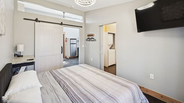 A bedroom with a bed, a desk with a lamp, a TV mounted on the wall, and a hallway leading to a kitchen.
