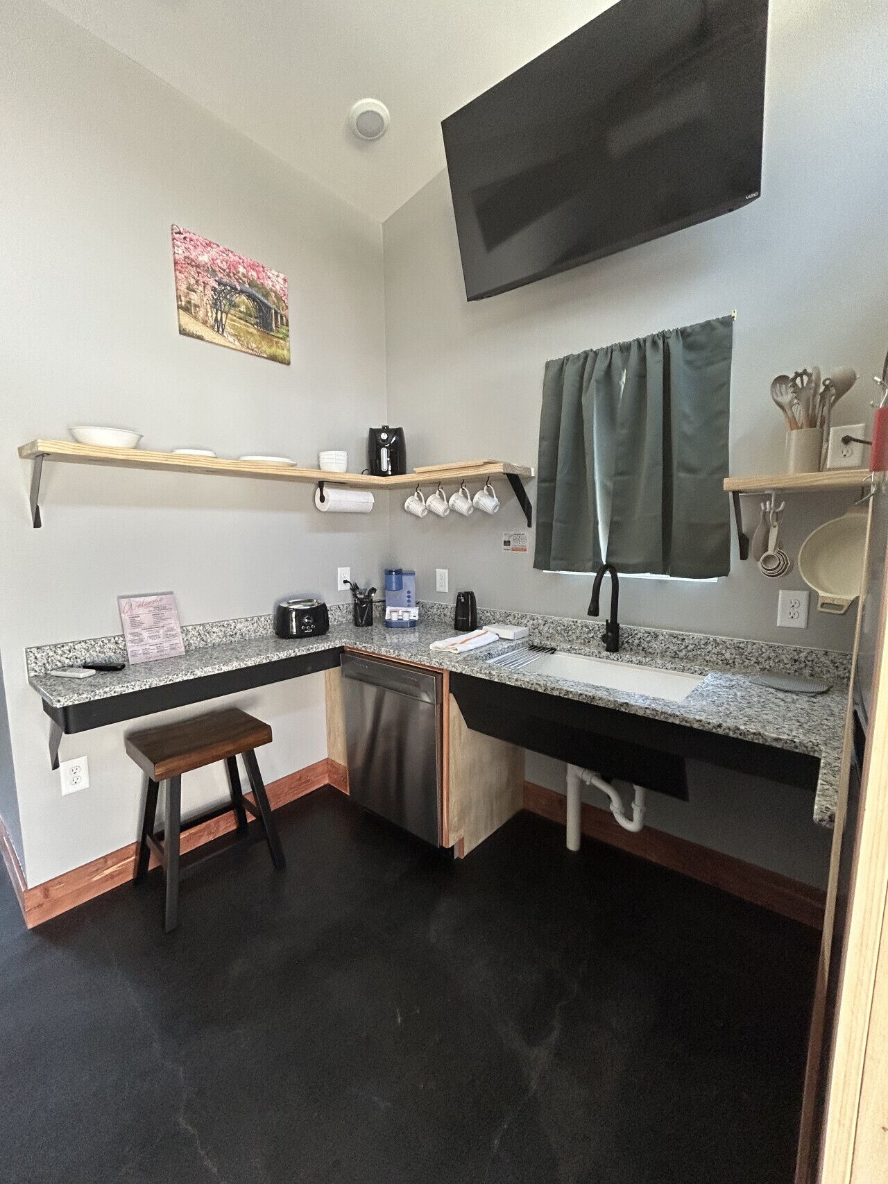 A compact kitchen with a built-in dishwasher, a small stool, a coffee maker, and a TV mounted on the wall.