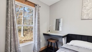 A bedroom with a bed, a nightstand, a window with curtains, a mirror, and a view of trees.