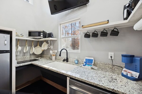 A kitchen with a granite countertop, stainless steel appliances, hanging pots, a window with a view of trees, and a mounted television.
