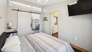 A bedroom with a bed, a desk with a lamp, a TV mounted on the wall, and a hallway leading to a kitchen. - Cabins off the Trace: Cabin 4 (Summertown)