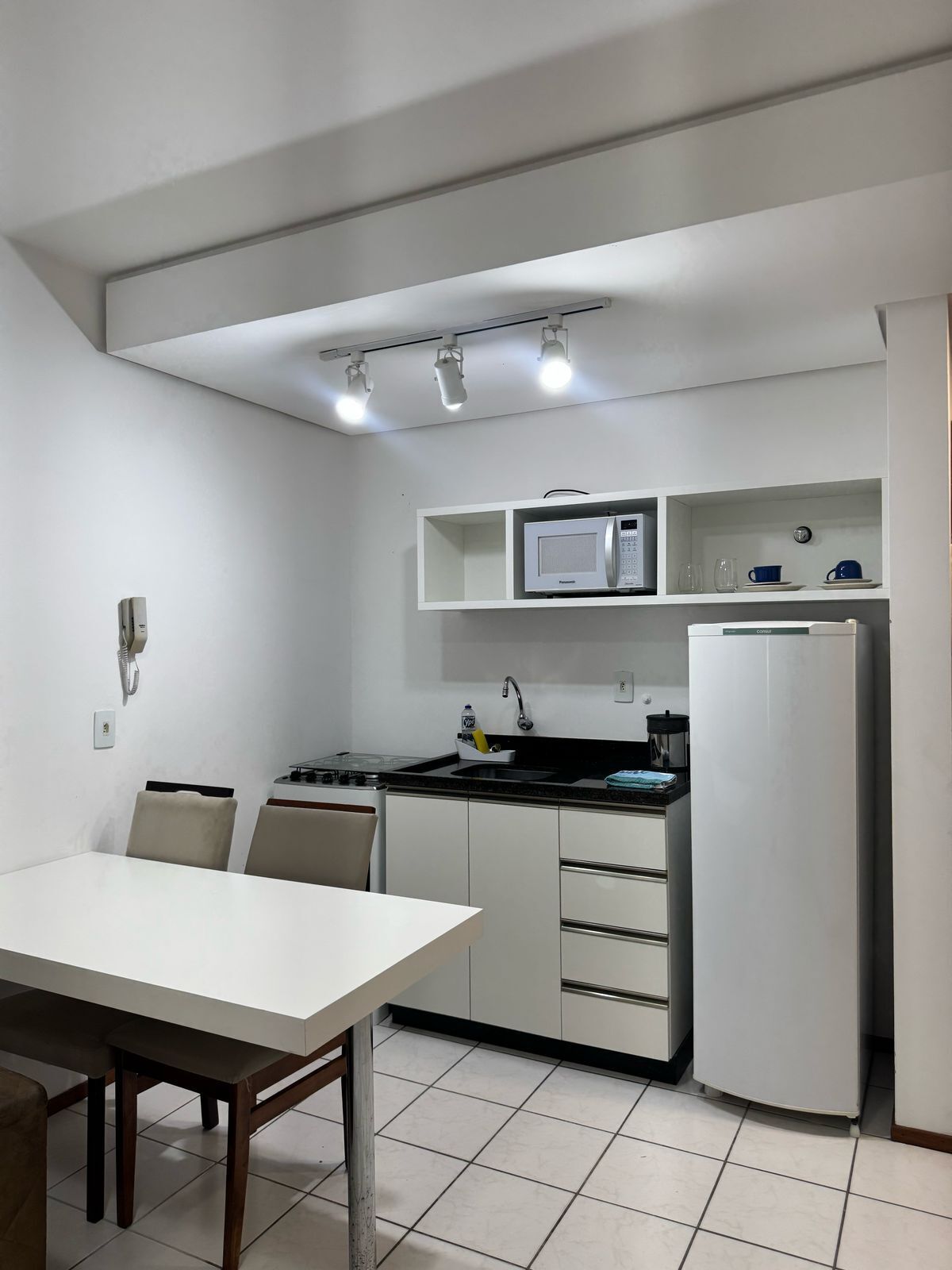 Cocina compacta con armarios blancos, un microondas, estufa y refrigerador. Hay una mesa de comedor con sillas y un pequeño estante sobre el microondas.