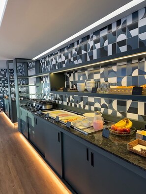 Un comptoir de cuisine moderne, doté d’un revêtement mural géométrique en carreaux, d’appareils électroménagers en acier inoxydable et offrant un assortiment de produits pour le petit-déjeuner, notamment des fruits et des viennoiseries.