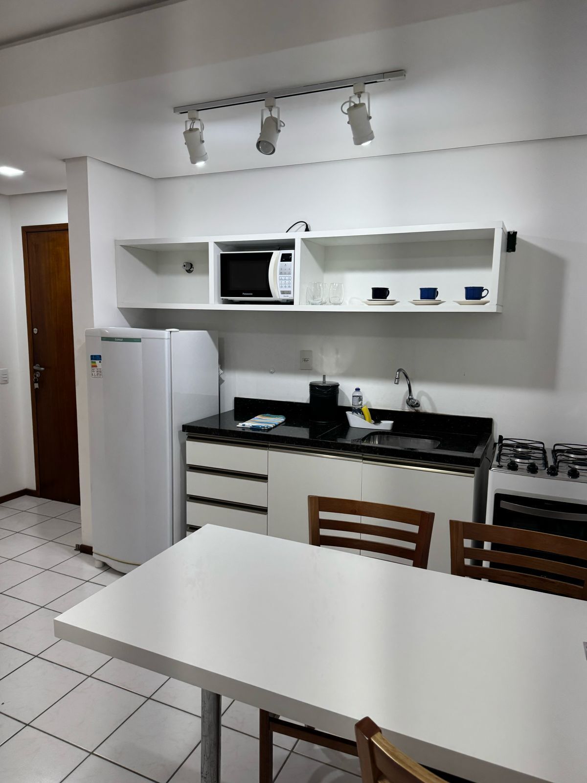 Cocina compacta con microondas, cafetera y cocina a gas. Hay una mesa blanca y dos sillas.