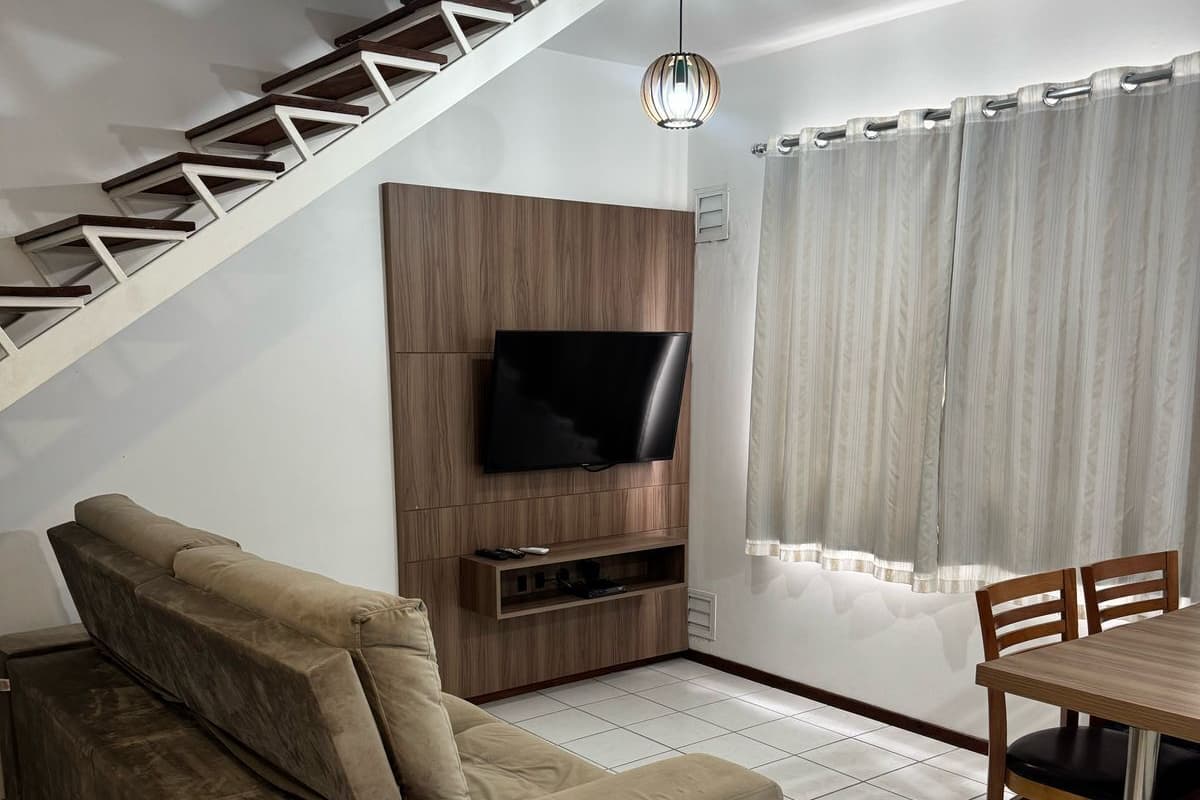 Een woonkamer met een bank, een aan een houten muur bevestigde televisie, een eettafel met stoelen en een modern vormgegeven trap.