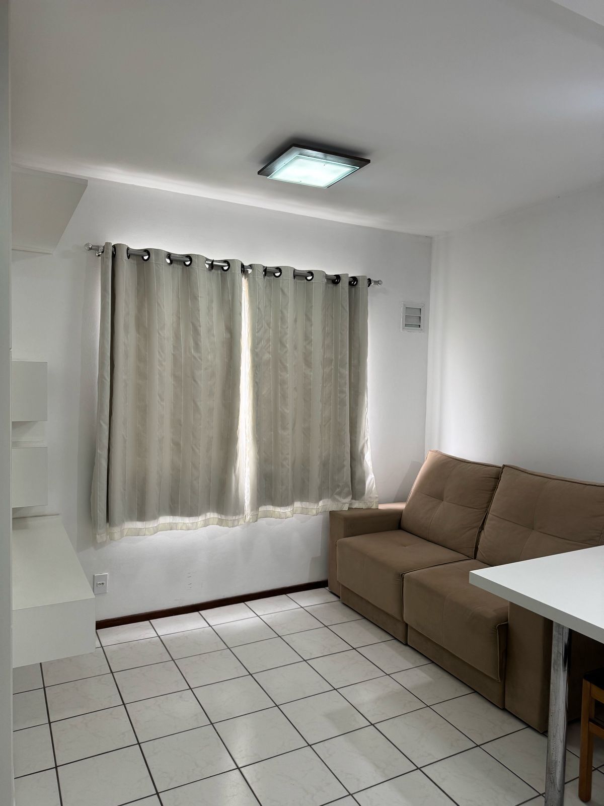 Um quarto com um sofá bege, uma mesa branca e uma janela com cortinas bege.