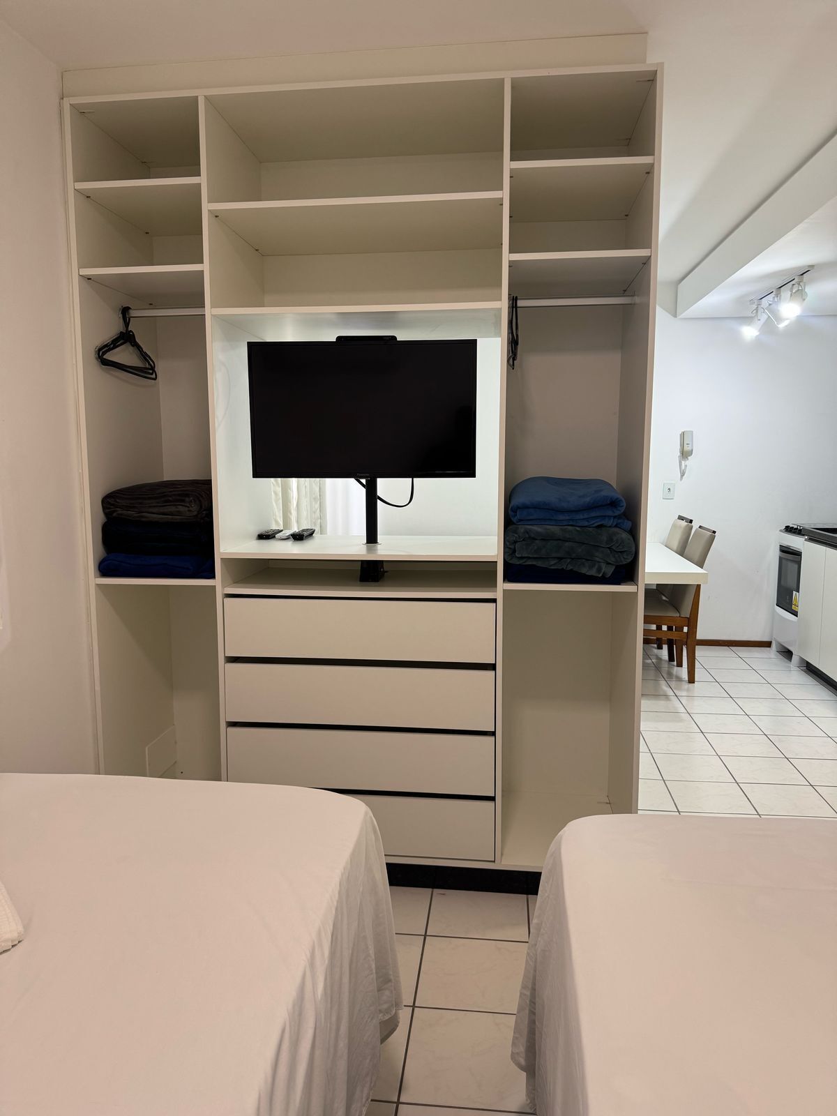 A bedroom with a TV on a stand, a wardrobe with multiple shelves, and a dining area with a table and chairs.