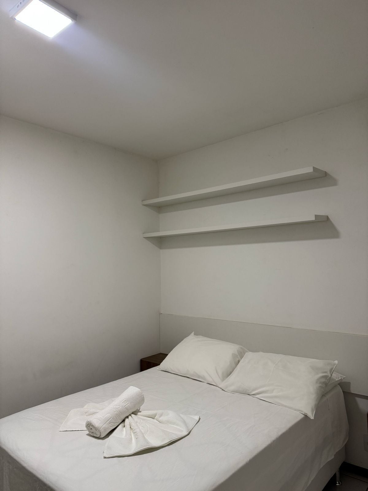 A simple, clean room with a bed, a nightstand, and two wall-mounted shelves.