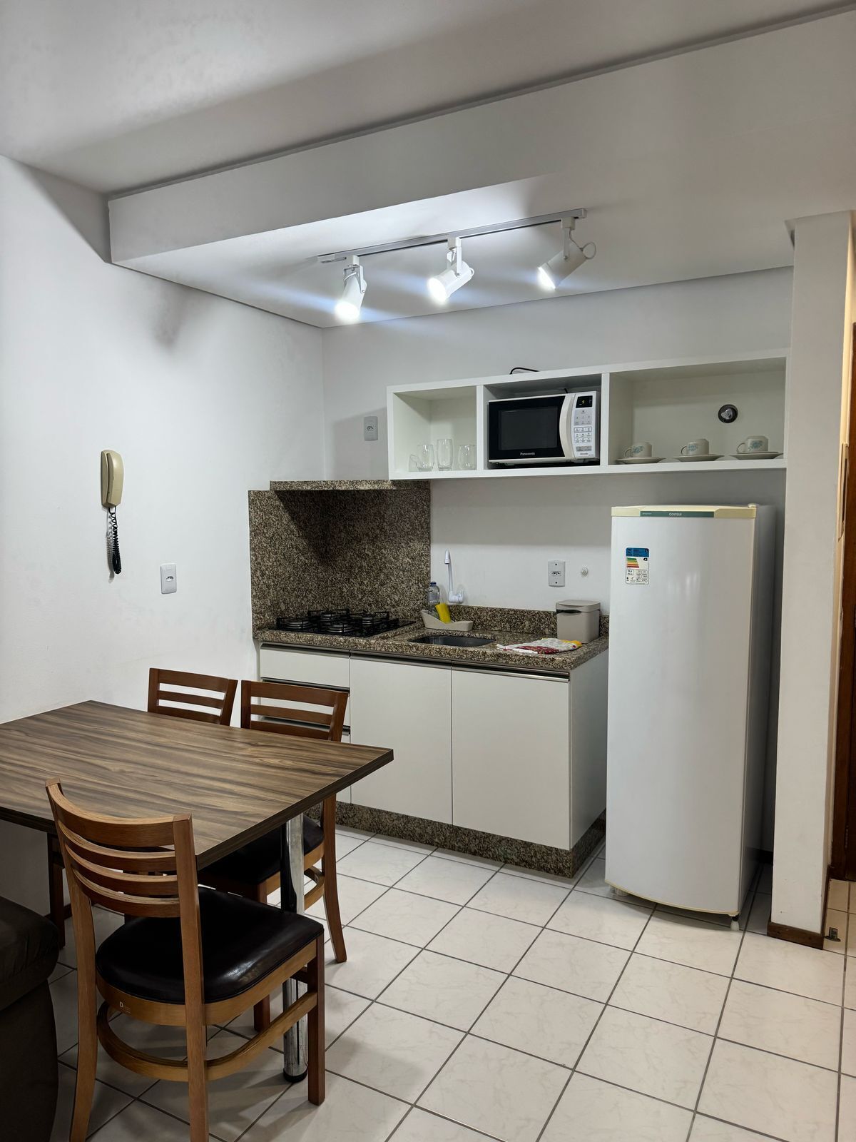 Una cocina compacta con armarios blancos, encimera de granito, microondas, refrigerador y una mesa de comedor con sillas.