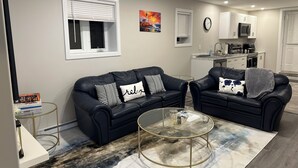 A modern living room with two black leather sofas, a glass coffee table, and a kitchen in the background.