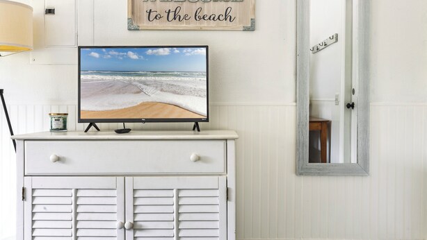 Un mueble blanco con un televisor que muestra una escena de playa, un cartel de bienvenida y un espejo que refleja una puerta.