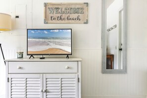 A white cabinet with a television displaying a beach scene, a welcome sign, and a mirror reflecting a door. - Peaceful Beach Bum – A Relaxed Butler Beach Retreat (St. Augustine)