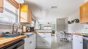 Una cocina moderna con encimeras de madera, armarios blancos, lavavajillas y un comedor con sillas blancas.