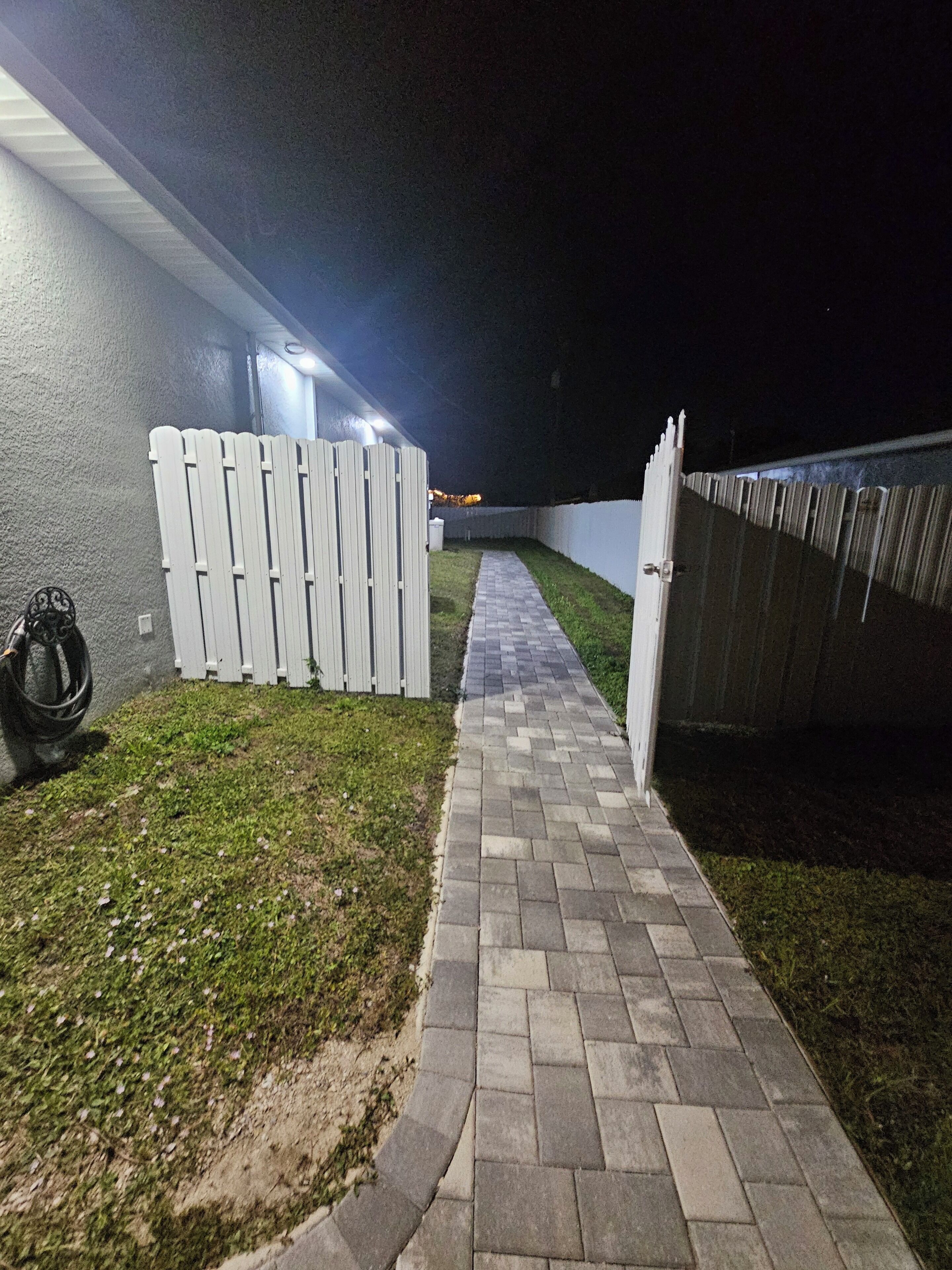 Un chemin pavé mène à une zone délimitée par une barrière et une clôture blanche, ainsi qu’un mur d’un côté.