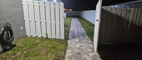 Un chemin pavé mène à une zone délimitée par une barrière et une clôture blanche, ainsi qu’un mur d’un côté.