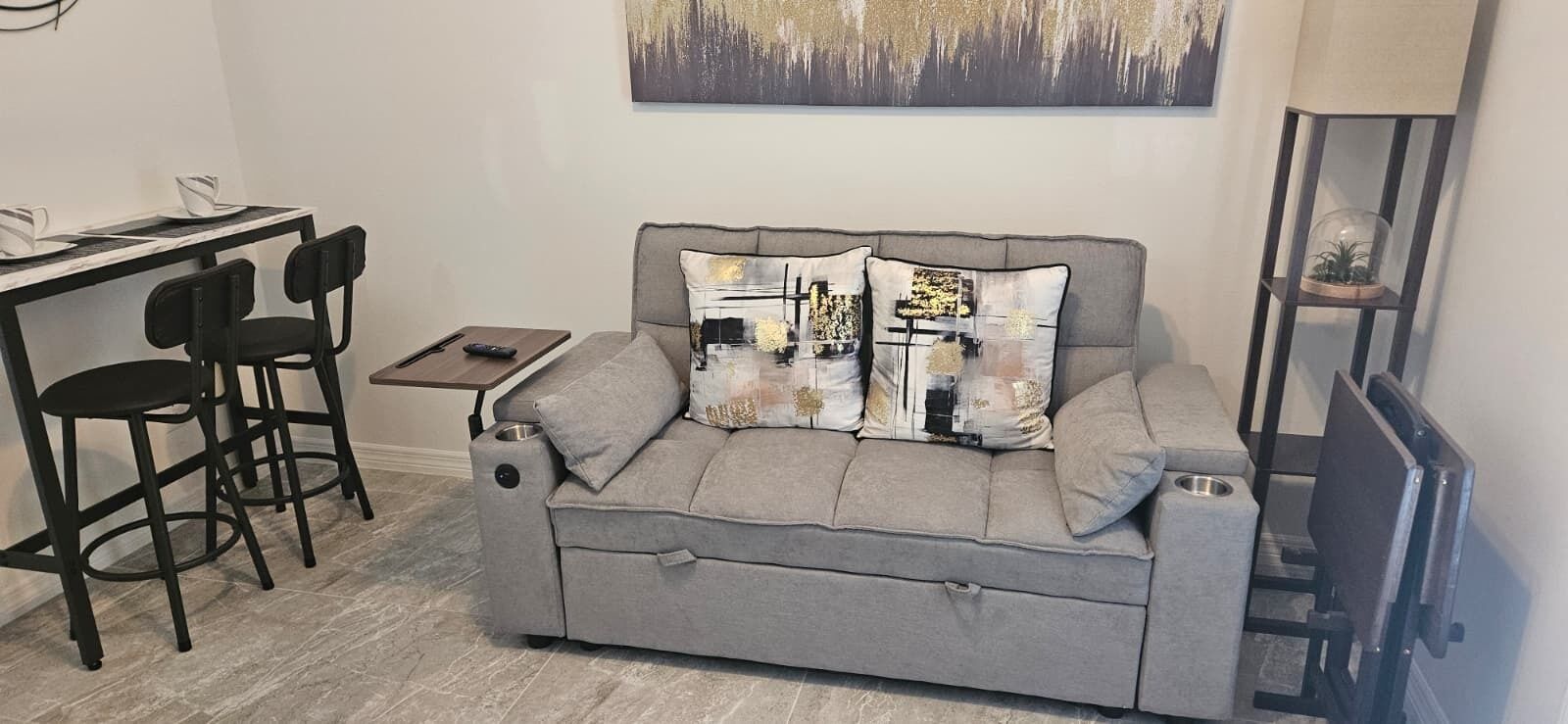 Un canapé gris agrémenté de coussins décoratifs, une petite table d’appoint et une chaise noire dans une pièce moderne ornée d’un tableau.
