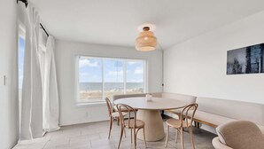 A room with a round table, four chairs, a bench, a hanging light fixture, a large window with a beach view, and a painting on the wall. - Oceanfront w/ Private Balcony | Quiet Views (North Myrtle Beach)