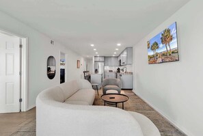 Ein modernes Wohnzimmer mit einer Couch, Sesseln und einem Couchtisch. Im Hintergrund ist eine Küche mit weißen Schränken und einem Essbereich zu sehen.